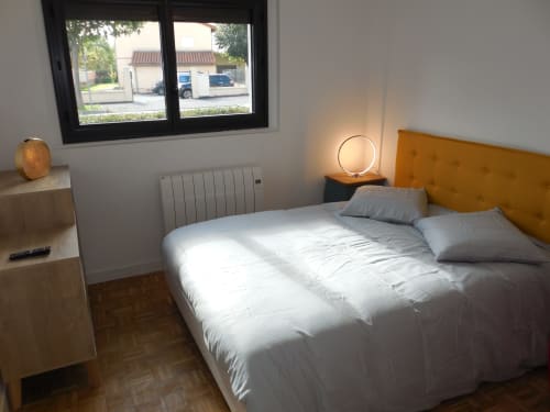 Chambre moderne dans un appartement toulousain avec literie blanche, tête de lit ocre, vue sur fenêtre et mobilier contemporain