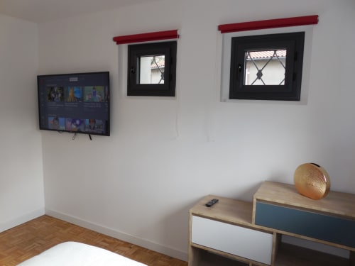 Chambre moderne à Toulouse avec TV murale, deux fenêtres à encadrements rouges, bureau en bois et panier de décoration tressé