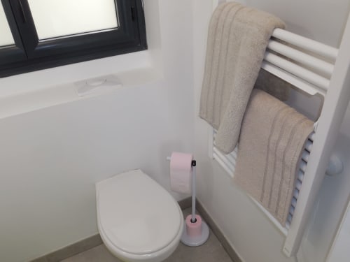 Salle de bain moderne à Room In Toulouse avec toilettes blanches, brosse WC rose, serviettes beige sur porte-serviettes chauffant et cadre noir