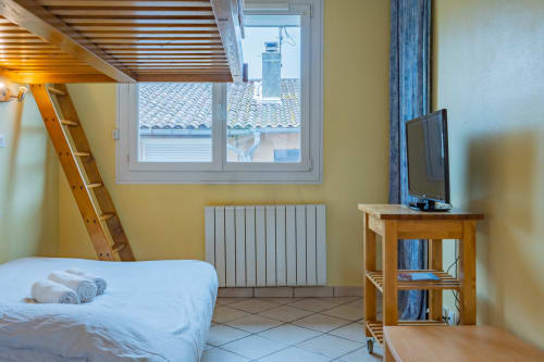 Chambre mansardée lumineuse dans un appartement toulousain avec plafond en bois, lit simple, télévision, radiateur et vue sur les toits de la ville