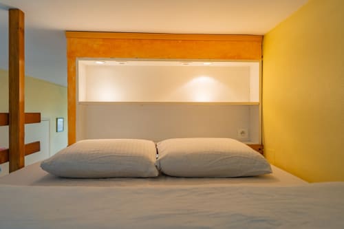Chambre moderne à Room In Toulouse avec deux oreillers, tête de lit en bois, mur jaune pastel et éclairage intégré à La