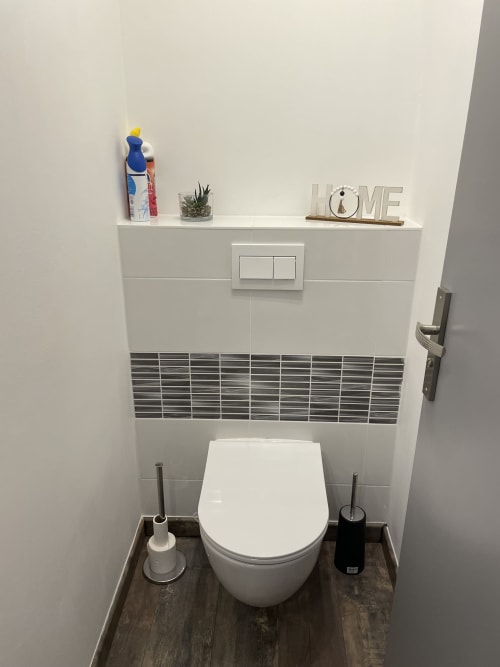 Salle de bain moderne avec toilettes blanches, accents de carrelage gris, étagère flottante décorée, et deux brosses de toilette chez Room In