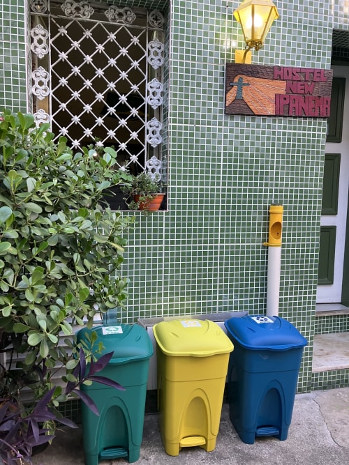 Entrada da Pousada New Ipanema com fachada de azulejos verdes, porta decorativa e três lixeiras de reciclagem coloridas no Rio de Janeiro