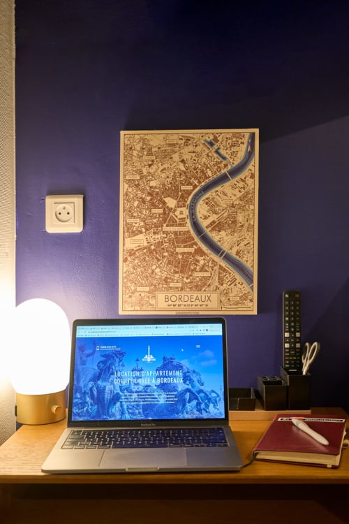 Desk workspace at Wellkhome Bordeaux with laptop displaying apartment rental site, wooden desk map of Bordeaux, lamp, and