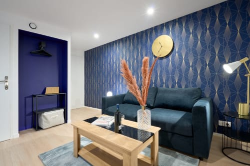 Modern studio apartment in Bordeaux with dark sofa, wooden table, blue patterned walls, and equipped kitchenette
