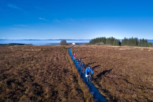 Cyclists ride along a blue path through moorland near Sliabh Beagh Hotel, Monaghan, with lake and forest views.