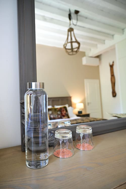 Water bottle and glasses on wooden desk in Pavillon Des Millesimes hotel room with mirrored view of bedroom