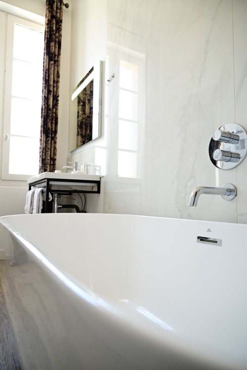Modern bathroom with white bathtub, illuminated mirror, marble walls, and city views at Pavillon Des Millesimes & SPA, Lussac