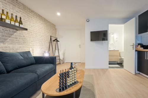 Modern studio apartment in Bordeaux with dark sofa, wooden table, brick accent wall, and open kitchen layout
