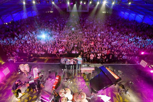 Large concert venue with thousands of spectators, colorful stage lighting, and live band performing on stage near Clarac,
