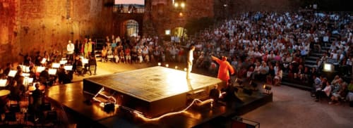 Grand theater event with orchestra and performers on illuminated stage before large audience in historic venue