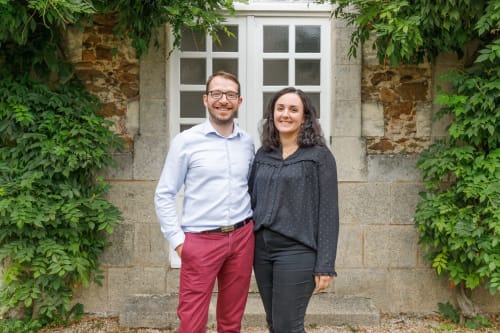 Deux hôtes souriants à La Maison D'olivier B&B à Mesnard la Barotière, France, devant l'entrée en pierre couverte de lierre
