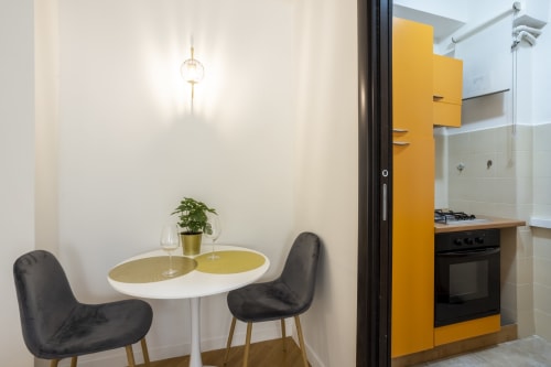 Zona pranzo moderna dell'hotel Re di Roma con tavolo rotondo, sedie nere, pianta in vaso e cucina gialla visibile a Roma
