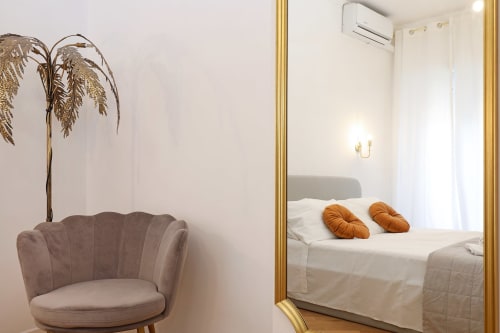 Camera da letto di un moderno appartamento romano con specchio cornice dorata che riflette il letto con cuscini arancioni, poltrona grigia e muri bianchi