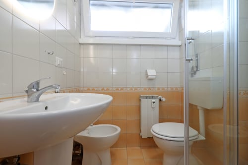 Bagno luminoso a Re di Roma con lavabo bianco, wc, doccia, dettagli in piastrelle arancioni e luce naturale dalla finestra