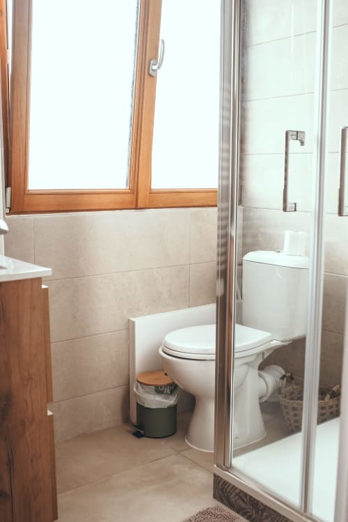 Salle de bain moderne aux murs en carrelage beige, toilettes blanches, cadre de fenêtre en bois et douche à l'hôtel ART DE VOYAGER