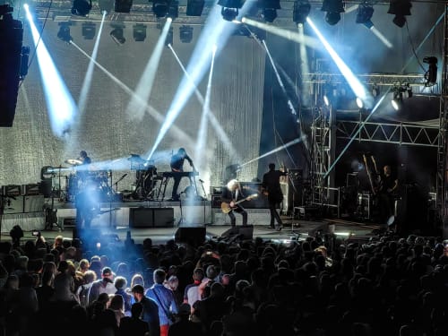 Live concert performance with band on stage, dramatic blue stage lighting, and crowded audience at Les Chalets Du Lac De