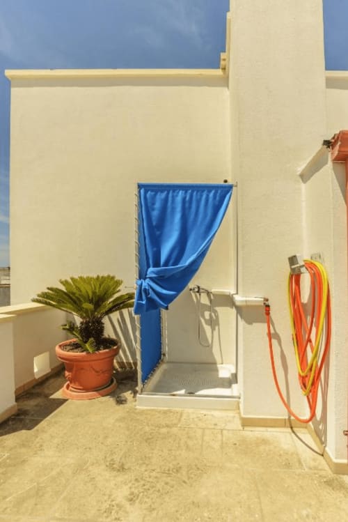 Area doccia esterna con tenda blu, palma in vaso e tubo arrotolato a Panoramici Monolocali a Leuca
