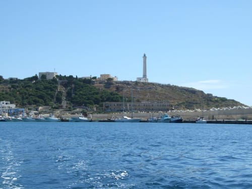 Porto costiero di Leuca con barche da pesca, faro sulla collina e acque blu del Mediterraneo