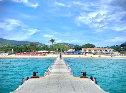 Wooden pier extending into turquoise Mediterranean waters at Villa Sigahi, La Croix Valmer, with beach and hillside buildings
