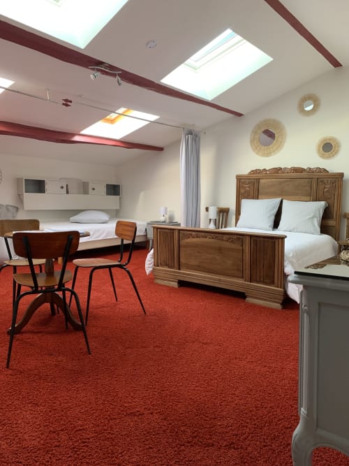 Grande chambre familiale Soleil sous les combles avec un grand lit en bois sculpté, deux lits simples, une table  avec des  chaises et une moquette rouge vif sous des fenêtres de toit.