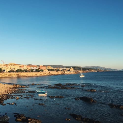 Veduta costiera di Alghero con edifici colorati, barche a vela e spiagge rocciose sotto un cielo azzurro sereno