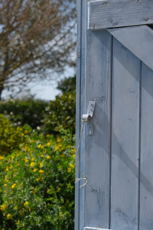 Weathered grey wooden door at Maison Monsieur Léger with rope pull and gate latch, garden flowers visible