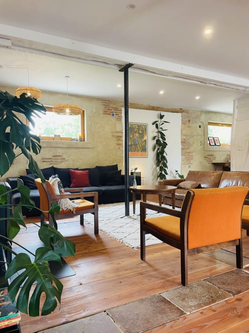Modern lounge at Maison Monsieur Léger with orange chairs, black sofa, wooden beams, and lush plants in Montaut.