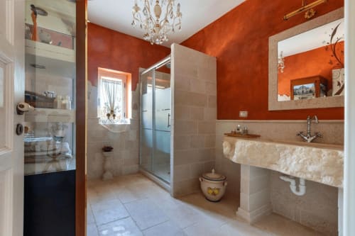 Elegante bagno a Villa Margherita con pareti color arancio bruciato, doccia in vetro, lavabo in pietra, lampadario e dettagli decorativi