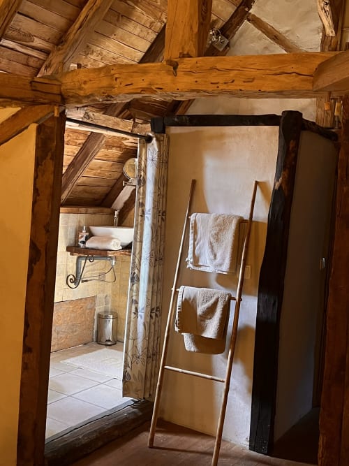 Chambre rustique aux poutres apparentes, échelle en bois chargée de serviettes, et porte ouverte sur la salle de bain au Lapin sur le Toit,