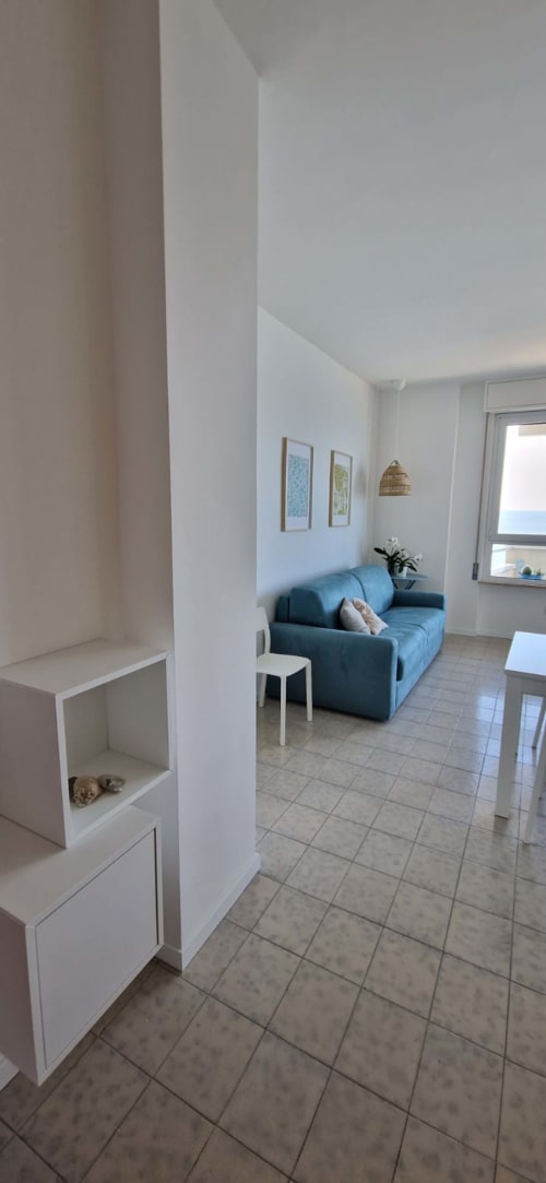Modern seafront apartment living room with blue sofa, white pillars, tile flooring, framed artwork, and sea view balcony at