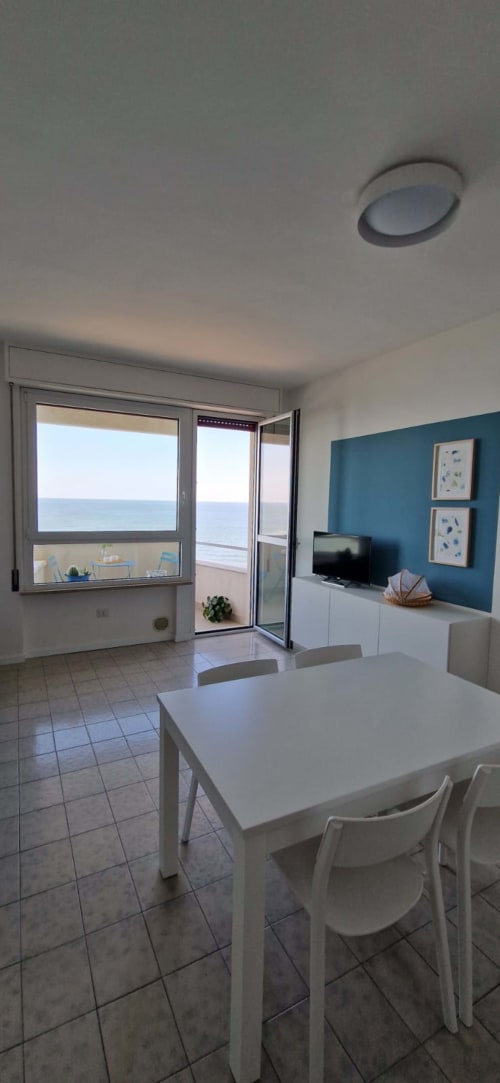 Seafront apartment dining area with sea view balcony, white table, teal accent wall, TV, and coastal window at Lamar Suites