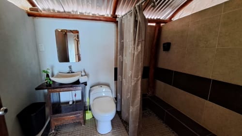 Baño moderno en el hostal Cayuco Maya de Bacalar con inodoro de cerámica blanca, tocador de madera, espejo y paredes alicatadas en tonos tierra