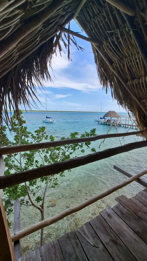 Vista desde habitación con techo de palapa en hostal Cayuco Maya, con vistas al lago turqueso de Bacalar, botes y muelle