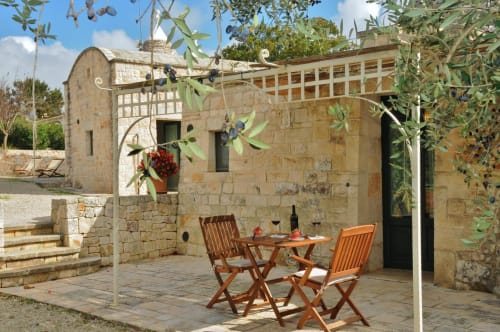 Cortile del Trulli Vulés a Ostuni con edificio in pietra, sedie in legno, tavolo da pranzo e ulivi che ombreggiano