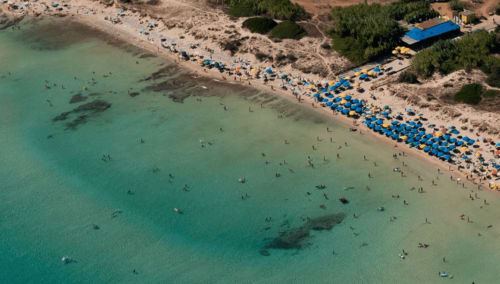Vista aerea della spiaggia di Leuca con acque turchesi, bagnanti e ombrelloni blu sulla riva sabbiosa vicino Panoramici Monolocali