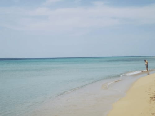 Spiaggia serena a Leuca con acque turchesi, lido sabbioso e una persona che passeggia nel mare cristallino