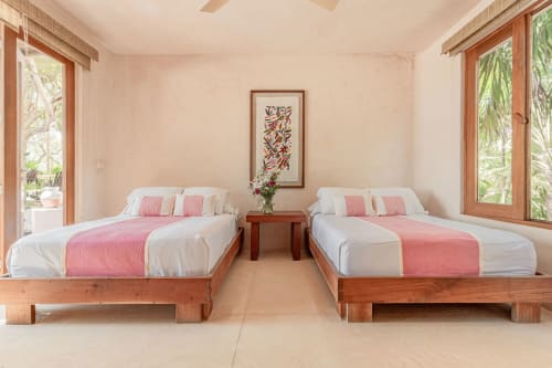Twin beds with pink accents in Cantarena House villa, Tulum. Wooden frames, ocean view doors, bright tropical setting.