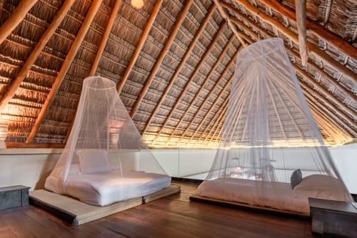 Spacious Tulum villa bedroom with sheer mosquito nets, wooden beams, and polished wood flooring under thatched roof