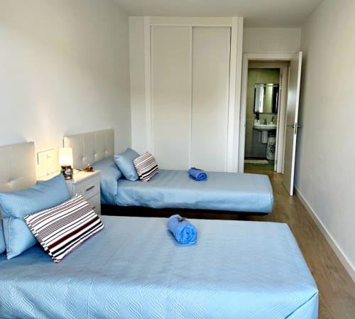 Moderno dormitorio en apartamento de Baiona con dos camas, ropa de cama azul, almohadas rayadas, lámpara de noche, armario empotrado y