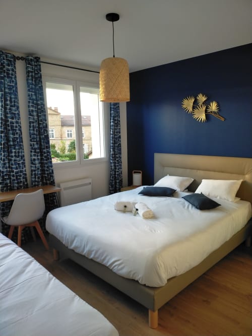 Chambre moderne au Gîte des Gônes avec lit beige, mur bleu marine, luminaire suspendu tissé, rideaux à motifs et jardin