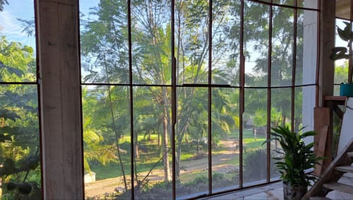Vistas al jardín desde la habitación del hostal Cayuco Maya en Bacalar, México, a través de grandes ventanales con árboles tropicales y áreas verdes visibles