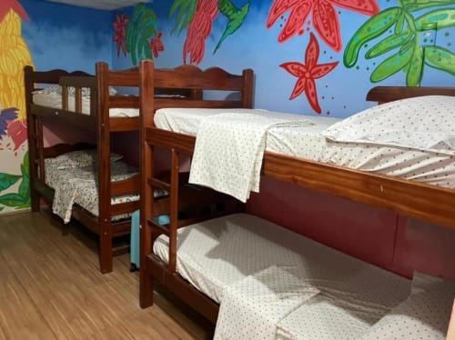 Quarto luminoso no Hostel New Ipanema Rio com beliches de madeira, mural tropical colorido e roupa de cama creme