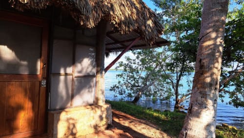Habitación rústica junto al lago en el hostel Cayuco Maya en Bacalar con techo de palma, puerta de madera y vista turquesa del lago entre árboles