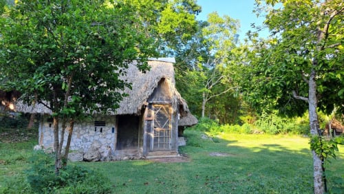 Cabaña de piedra rústica con techo de paja rodeada de vegetación exuberante en el hostal Cayuco Maya de Bacalar, México