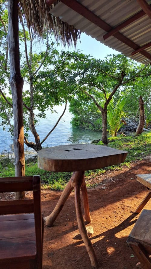 Mesa de madera redonda bajo techo de palapa con vista al lago Bacalar y mangles en el hostal Cayuco Maya