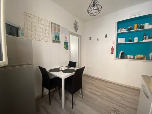 Zona pranzo di moderno appartamento a Bari con tavolo bianco, sedie nere, scaffali turchesi e servizi cucina