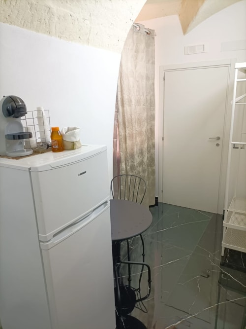 Camera compatta del Newhouse Bari con angolo cottura, minifrigo, sedia da pranzo e tende beige