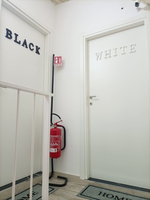 Corridoio dell'hotel Newhouse a Bari con porte bianche e nere etichettate, estintore e segnaletica di emergenza