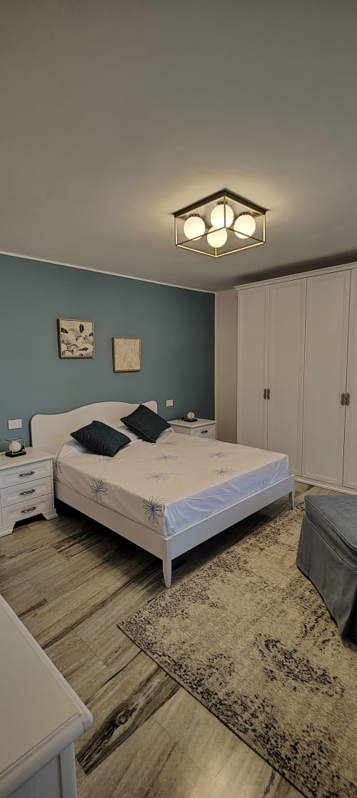 Camera da letto moderna con parete grigio-blu, letto bianco, armadio color crema, lampadario geometrico e pavimento in legno